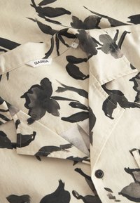 Beige Bluse mit floralen Mustern, schwarzen Aquarellblumen, einem Kragen und einem Knopfverschluss. Der Stoff wirkt leicht und hat eine glatte Textur.