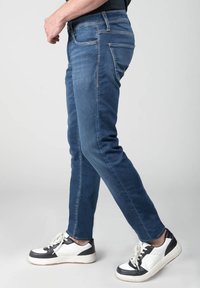 Vaqueros de mezclilla azul de corte ajustado con costuras amarillas en contraste, cinco bolsillos, cintura de altura media, combinados con zapatillas en blanco y negro.