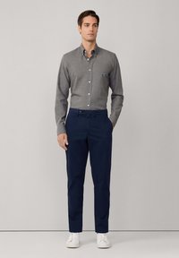 Hombre de pie con las manos en los bolsillos, vistiendo una camisa de botones de manga larga gris, pantalones navy y zapatillas blancas frente a un fondo liso.
