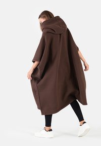 Brązowy oversize'owy poncho z kapturem, wykonane z miękkiego polaru, z dużym kapturem, krótkimi rękawami i luźnym krojem, w połączeniu z czarnymi legginsami.