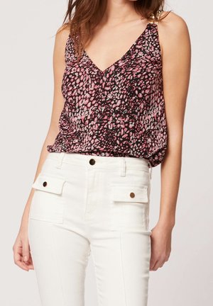 Frau trägt ein ärmelloses V-Ausschnitt-Top mit rosa-schwarzem Muster und eine hoch taillierte weiße Hose mit vorderen Klappentaschen und Knöpfen.