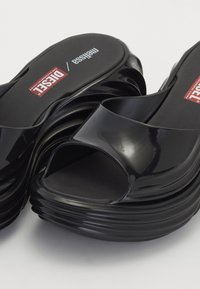 Sandale platform din plastic negru, cu un design cu vârful deschis. Textură în dungi pe talpă și branding proeminent pe brant.