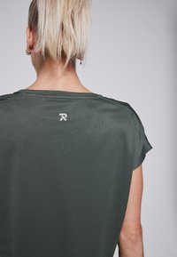 Dark green ribbed short-sleeve top with a rounded neckline, featuring a small white logo on the back. Soft texture and relaxed fit.