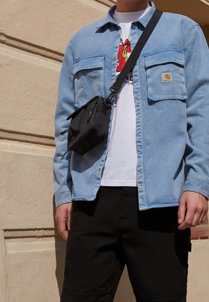 Person wearing a light blue button-up jacket over a white graphic t-shirt, black pants, and a black crossbody bag.