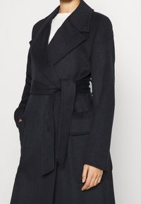 Donkergrauwe wollen jas met brede revers, een strikceintuur in de taille, klepzakken, gedragen over een wit shirt.