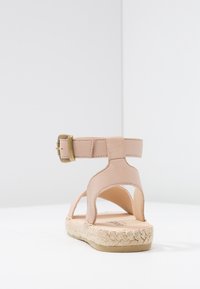Beige lädersandal med en spänne vid vristen och öppen design, med en espadrille-liknande sula av jute och slät yta.