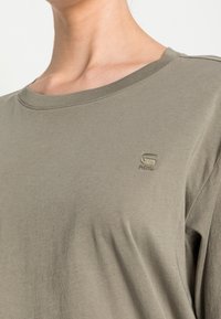 Närbild av en person som bär en dämpad grön T-shirt med rund hals, med en liten broderad logotyp på bröstet som säger "RAW."