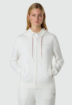 Giovane donna che indossa una felpa bianca con zip e coulisse rosa chiaro, e pantaloni bianchi abbinati, in piedi con le mani nelle tasche.