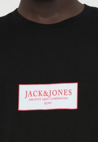 Μαύρη μπλούζα φτιαγμένη από βαμβάκι, με λευκό ορθογώνιο λογότυπο και κόκκινο κείμενο που λέει "JACK & JONES" και "CREATIVE DEPT. COPENHAGEN."