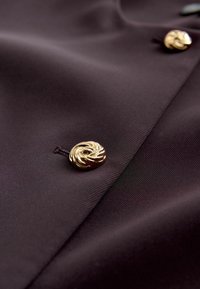 TumeLilla kangas, sileda tekstuuriga, millel on silmatorkav kuldse spiraaliga nööp. Lihtne, elegantne disain nähtavate õmblustega.