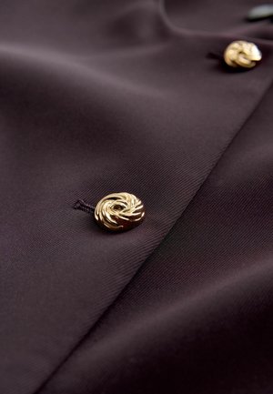 Ciemnofioletowa tkanina o gładkiej fakturze, z wyraźnym złotym guzikiem w kształcie spirali. Prosty, elegancki wzór z widocznym przeszyciem.