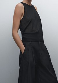 Top noir sans manches avec une texture lisse et une taille cintrée, associé à un pantalon noir ample avec des plis et des poches latérales.