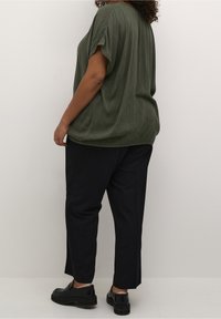 Løs, olivengrøn bluse med faldende skuldre, parret med sorte bukser og sorte slip-on sko, vist fra ryggen mod en neutral baggrund.