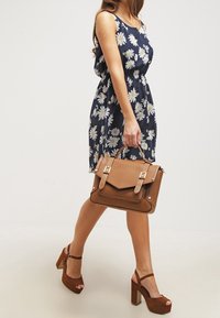 Hnědá kožená kabelka s klopou, zlatými kovovými detaily a dvěma horními uchy. Modelka má na sobě tmavě modré květinové šaty a hnědé platformové podpatky.