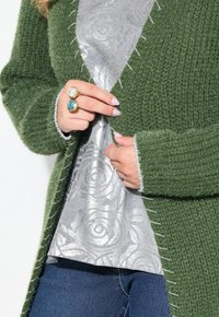 Grön stickad cardigan med texturerad garn, kontrasterande ljus sömnad, lager över en silverblommig topp. Händer visar ringar med olika stenar.