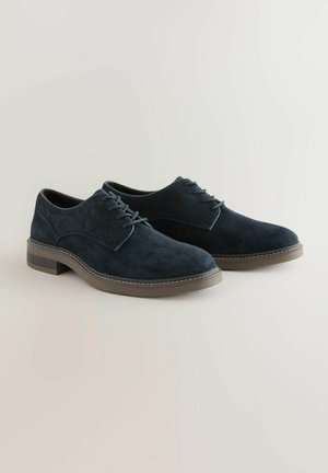 Une paire de chaussures de ville en daim bleu marine à lacets avec des semelles en caoutchouc marron sur un fond clair et neutre.