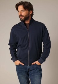 Marine blauwe zip-up jack van zachte stof met een hoge kraag, voorzien van zijzakken en een zipzak op de linkerarm, gecombineerd met een blauwe spijkerbroek.