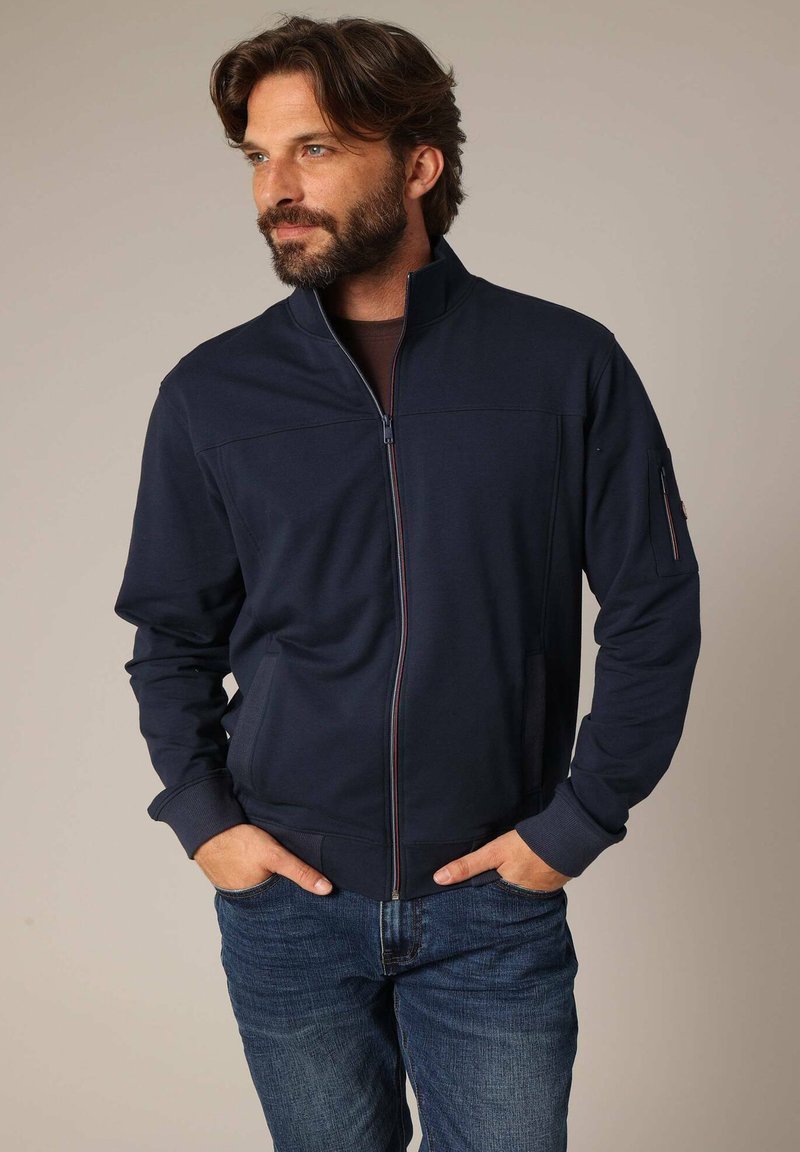 Marine blauwe zip-up jack van zachte stof met een hoge kraag, voorzien van zijzakken en een zipzak op de linkerarm, gecombineerd met een blauwe spijkerbroek.