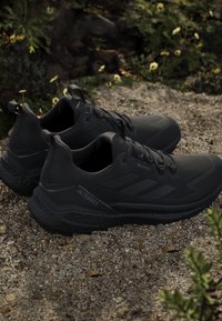 Scarpe da trail running Adidas Terrex nere posizionate sulla ghiaia con piante verdi e piccoli fiori gialli sullo sfondo.