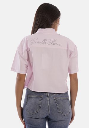 Donna con lunghi capelli scuri che indossa una maglietta rosa chiaro a maniche corte, larga, con la scritta "Guett Paris" sulla schiena e jeans di denim blu.