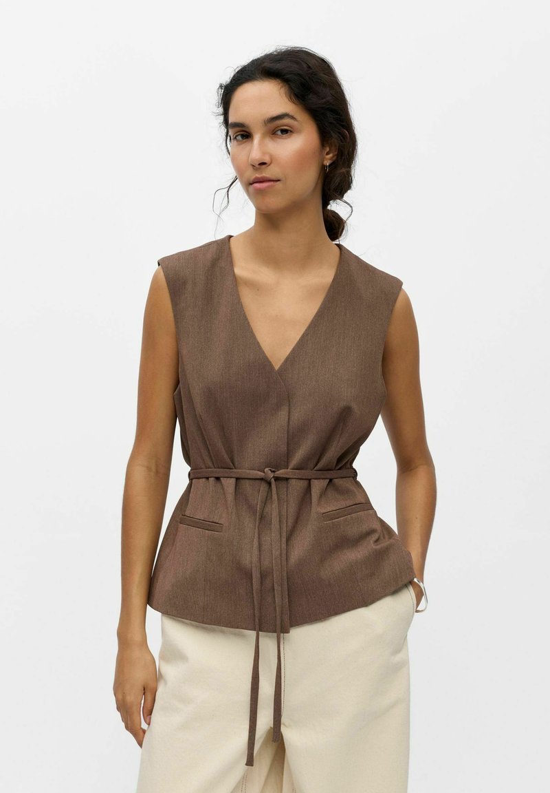 Femme aux cheveux foncés attachés, portant un gilet marron sans manches avec un lien à la taille et un pantalon clair sur un fond uni.