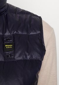 Granatowa kamizelka puchowa o błyszczącej, pikowanej teksturze, z czarną metką z żółtym napisem. Zapięcie na zatrzaski; noszona na jasnym swetrze.