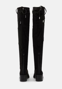 Botas altas de ante negro con cremalleras en la parte trasera, tacones de bloque y cordones atados en la parte superior, mostradas desde la parte trasera sobre un fondo liso.