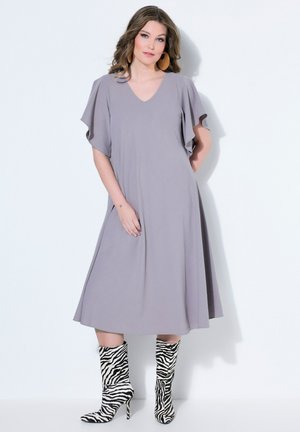 Vrouw draagt een loszittende lavendel midi-jurk met wapperende mouwen en hoge hakken met zebraprint tegen een witte achtergrond.