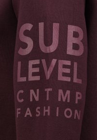 Bordeaux sweatshirt met grote, contrasterende gedrukte tekst: "SUB LEVEL CNTMP FASHION" in lichtpaars. Zachte stoftextuur.