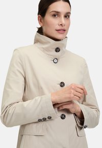 Beiger, langärmeliger Mantel mit hohem Kragen und schwarzen Knopfakzenten, der eine taillierte Silhouette und seitliche Taschen aufweist. Glatte Stofftextur.