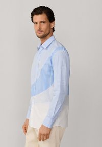 Man met kort bruin haar, gekleed in een overhemd met lange mouwen met blauw-witte verticale strepen en beige broek, staand zijwaarts.