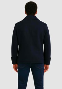 Giacca in misto lana blu navy con collo classico, vestibilità sartoriale e dettagli di cuciture visibili, abbinata a pantaloni in denim scuro.