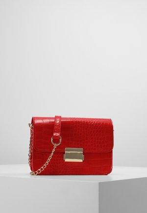 Rode schoudertas van krokodillenleer met een gouden kettingriem en een rechthoekige gouden sluiting, geplaatst op een wit voetstuk tegen een eenvoudige achtergrond.