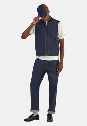 Uomo che indossa un gilet e pantaloni di denim blu scuro, t-shirt bianca, mocassini neri, calzini bianchi e un cappellino bicolore blu e marrone, che sistema la visiera del cappellino.