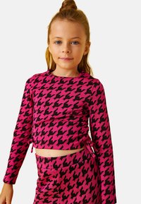 Niña pequeña con cabello castaño claro recogido en un moño, vestida con una blusa de manga larga de cuadros en rosa y negro y pantalones a juego.