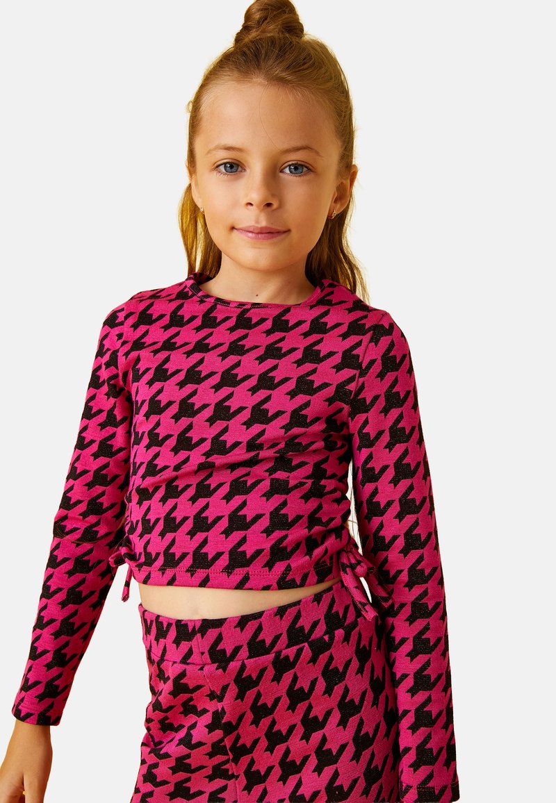 Niña pequeña con cabello castaño claro recogido en un moño, vestida con una blusa de manga larga de cuadros en rosa y negro y pantalones a juego.