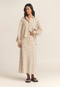 Beige polka dot shirt and matching skirt set, featuring a relaxed fit, button closure, and long sleeves. Worn with black slide sandals.