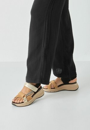Wedge sandals with a mix of beige, gold, and animal print straps, featuring a cushioned sole and textured black fabric pants.