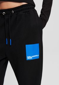 Schwarze Jogginghose mit einem Kordelzugbund, ausgestattet mit einem blauen rechteckigen Patch mit dem Text "KARL LAGERFELD JEANS" am linken Oberschenkel.