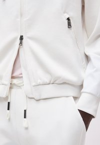 Λευκό μπουφάν με φερμουάρ μπροστά και τσεπάκι στο πλάι με φερμουάρ, με λευκή φόρμα με κορδόνι στο ίδιο χρώμα, χέρι σε τσέπη ορατό σε σκουρόχρωμο δέρμα.
