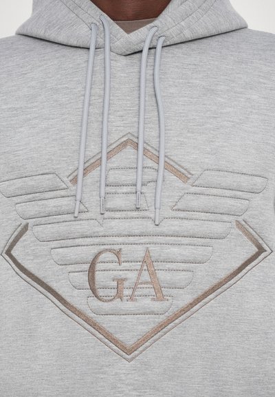 Grijze hoodie met een gestructureerd logo in brons, met een diamantvorm en vleugelachtige accenten. Inclusief lichtgrijze trekkoorden.