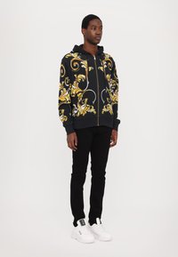 Felpa nera con zip e motivo ornamentale oro e bianco, abbinata a pantaloni neri e sneaker bianche. La felpa presenta una zip frontale e lacci.