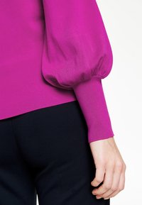 Fuchsia, ruggad tröja med puffärm och åtsittande ärmar, kombinerad med mörka byxor. Tyget verkar slätt och något glänsande.