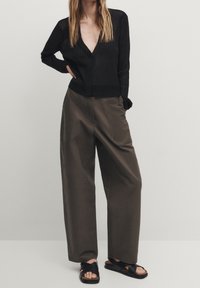 Schwarzer, gerippter Longsleeve-Pullover mit tiefem V-Ausschnitt, kombiniert mit hochgeschnittenen, lockeren olivgrünen Hosen und schwarzen Sandalen mit Riemchen.