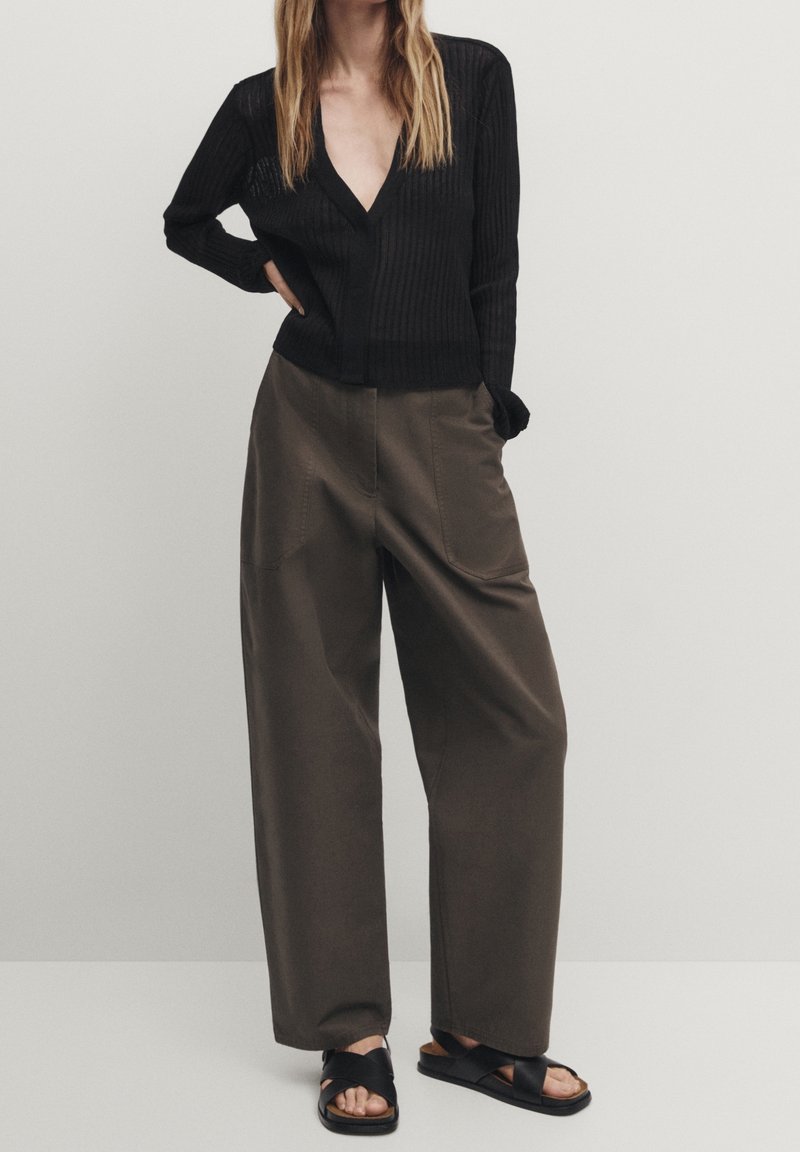 Schwarzer, gerippter Longsleeve-Pullover mit tiefem V-Ausschnitt, kombiniert mit hochgeschnittenen, lockeren olivgrünen Hosen und schwarzen Sandalen mit Riemchen.