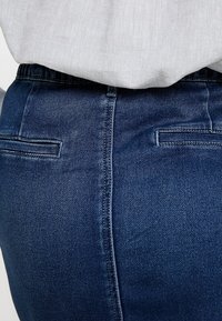Vaqueros de mezclilla azul oscuro con una textura suave, que presentan una cinturilla de altura media, corte recto y dos bolsillos traseros con detalles de costura mínimos.