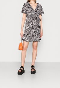 Robe portefeuille florale en tissu sombre, manches courtes, ourlet volants, associée à des sandales à plateforme noires et un petit sac à main orange.