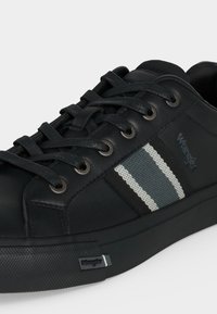 Baskets en cuir noir avec lacets noirs, détail en tissu rayé gris et blanc et logo Wrangler sur le côté et la semelle.