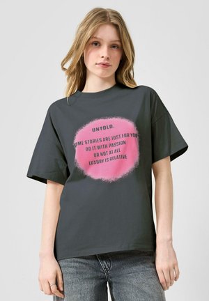 Junge Frau mit langen blonden Haaren, die ein dunkelgraues T-Shirt mit einem pinken Kreis und schwarzem Text trägt, vor einem hellgrauen Hintergrund stehend.