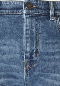 Gros plan de la ceinture d'un jean en denim bleu avec bouton métallique, passants de ceinture et détails de couture visibles.